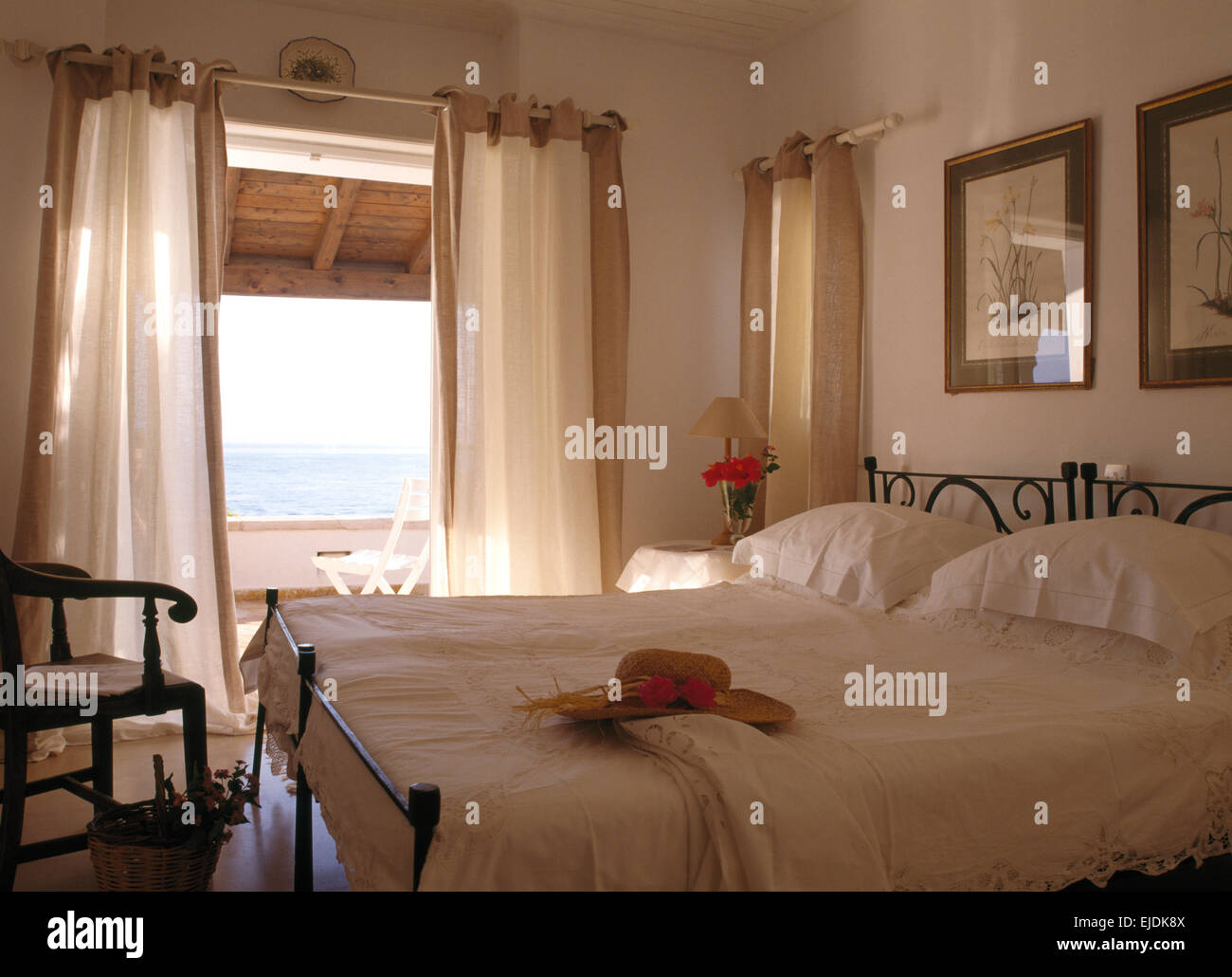 Weiße Wäsche auf Betten aus Schmiedeeisen in küstennahen Schlafzimmer mit weißen Vorhängen an Türen zum Balkon mit Blick auf das Meer Stockfoto