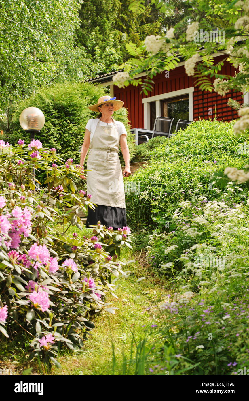 Ältere Frau, die in ihrem Garten, sie ist umgeben von Büschen. Es ist ein rotes Haus, teilweise im Hintergrund zu sehen. Sie ist weari Stockfoto
