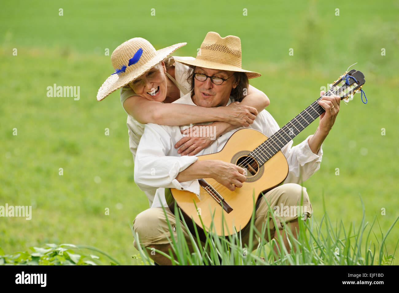 Älteres Paar gemeinsam Spaß haben. Der Mann sitzt und eine Gitarre zu spielen. Die Frau umarmt ihn von hinten. Sie sind im Freien. Stockfoto