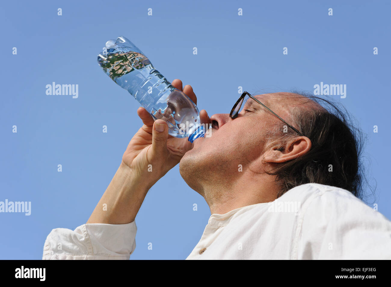 Durstig senior Mann trinkt Wasser aus der Flasche im Freien. Gibt es einen See und Wald im Hintergrund. Niedrige Kamerawinkel. Stockfoto