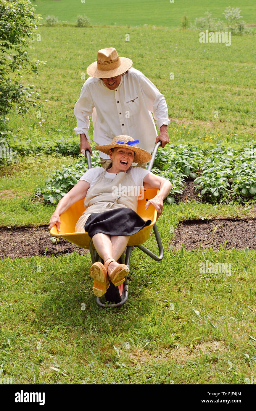 Ältere Mann trägt eine ältere Frau in einer Schubkarre im Freien in der Nähe ein Gemüsebeet. Sie lachen und Spaß haben. Stockfoto