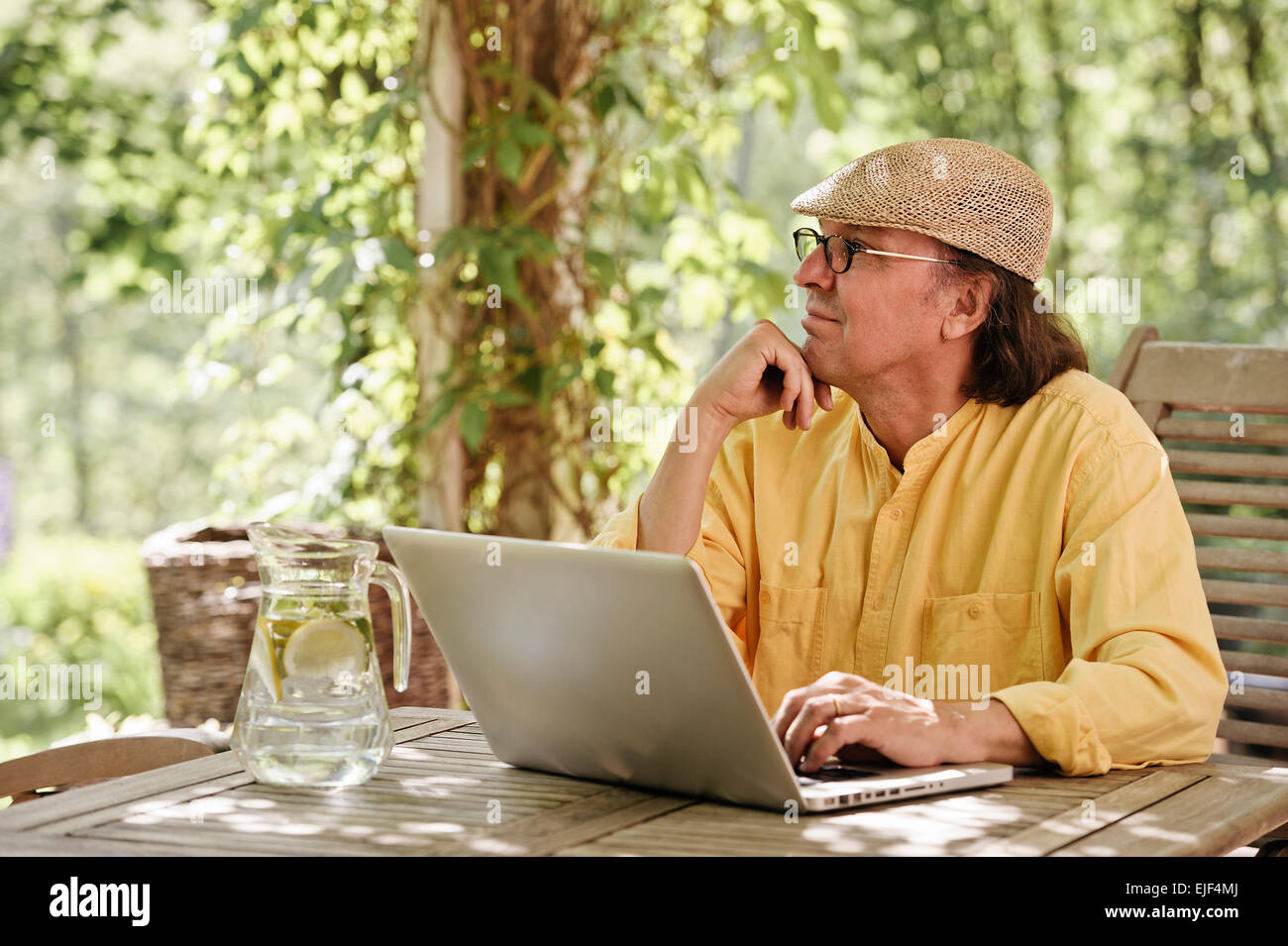 Ältere Mann sitzt im Freien an einem Holztisch unter einer Pergola und funktioniert auf einem Laptopcomputer. Es ist Sommer und es gibt ein Hintergrund Stockfoto