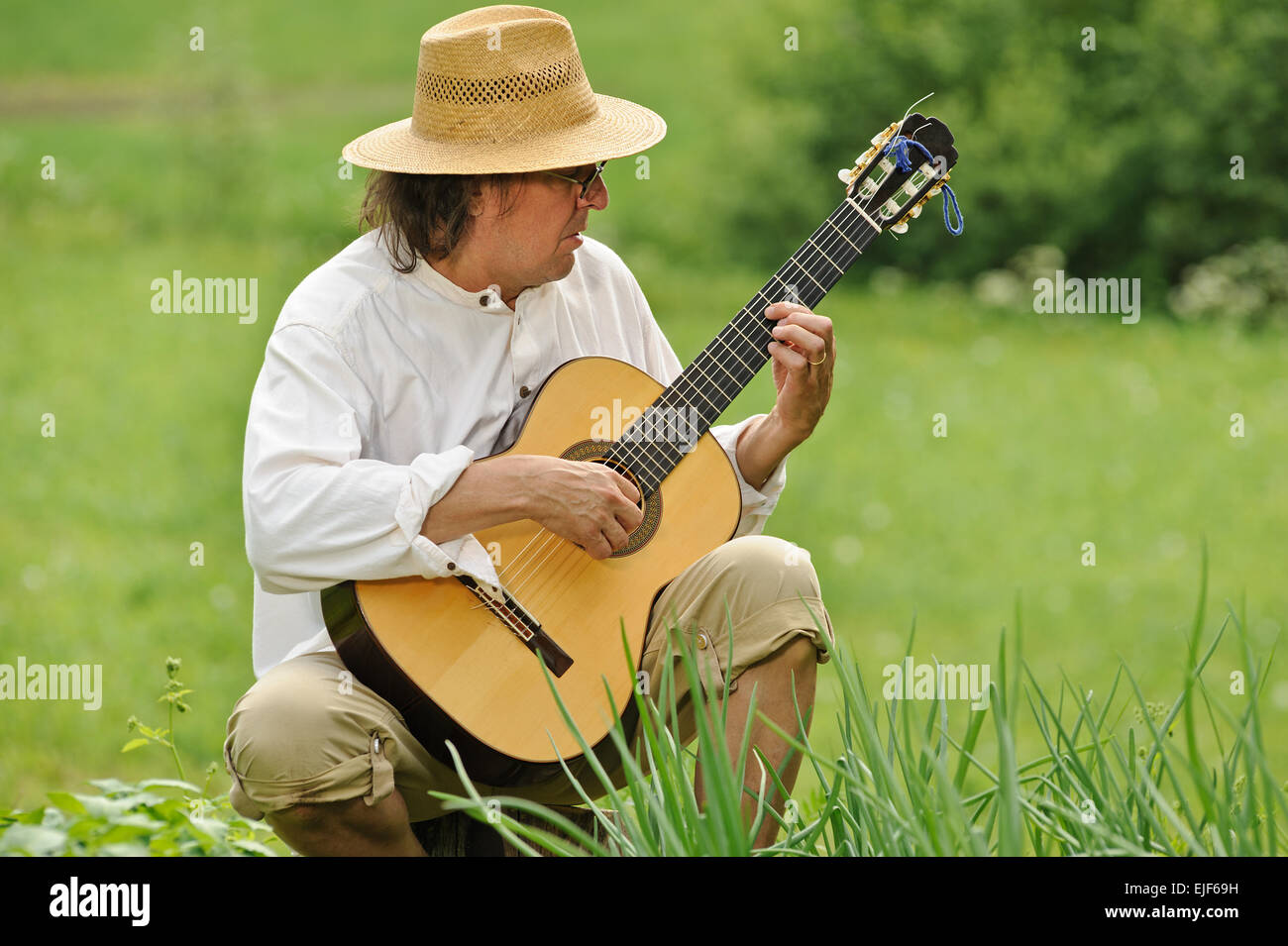 Senior Woman spielt eine Nylon-Saiten-Gitarre im Freien, er sitzt auf einem Baumstamm. Es gibt ein verschwommenes Feld im Hintergrund Stockfoto