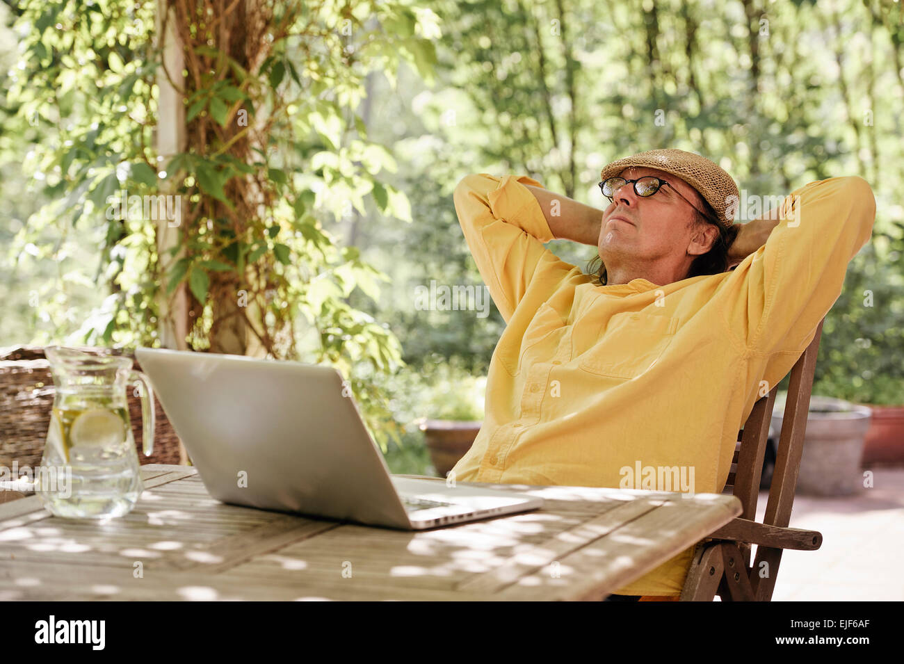 Ältere Mann sitzt im Freien an einem Holztisch unter einer Pergola und funktioniert auf einem Laptopcomputer. Es ist Sommer und es gibt ein Hintergrund Stockfoto