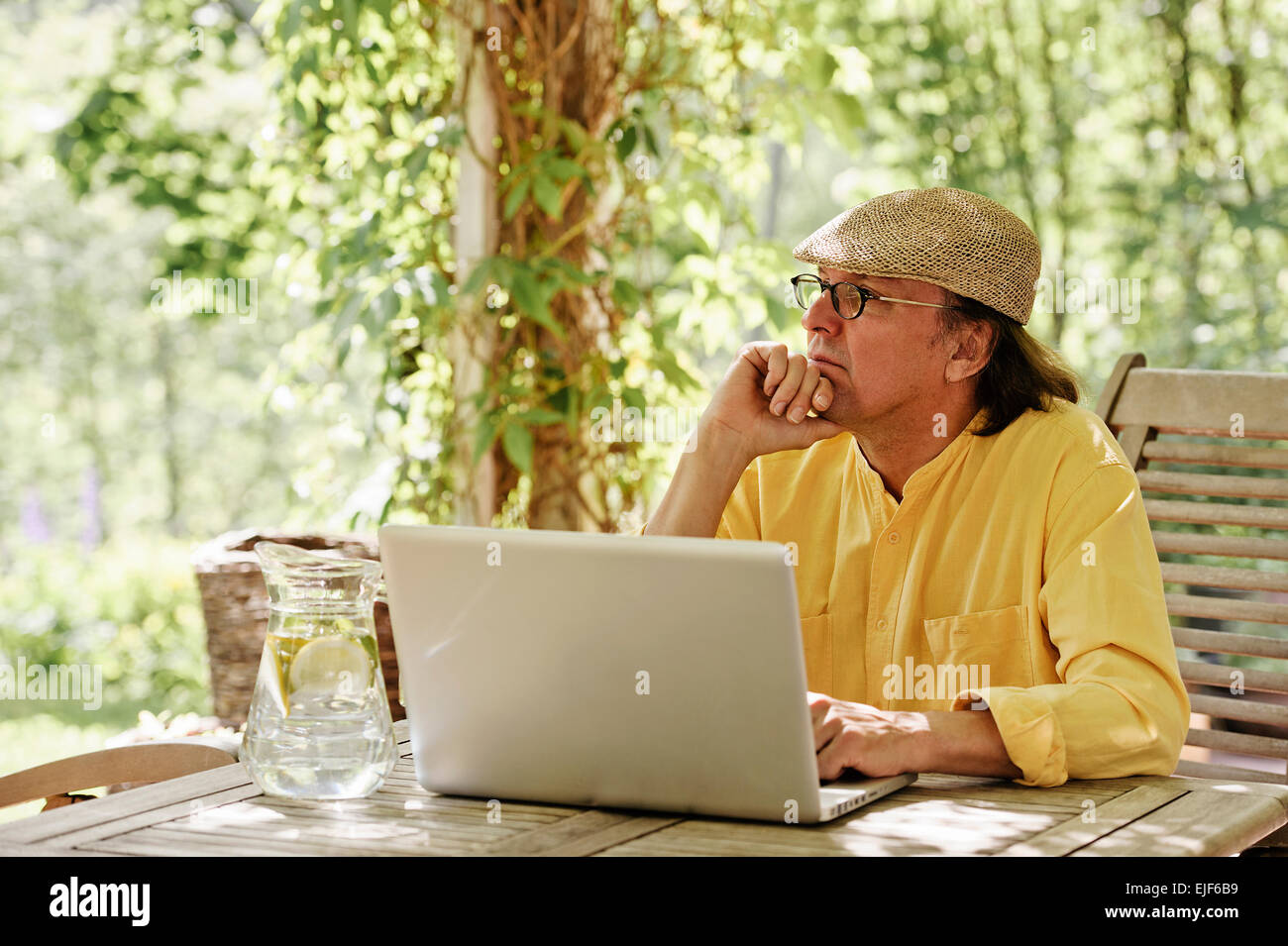 Ältere Mann sitzt im Freien an einem Holztisch unter einer Pergola und funktioniert auf einem Laptopcomputer. Es ist Sommer und es gibt ein Hintergrund Stockfoto
