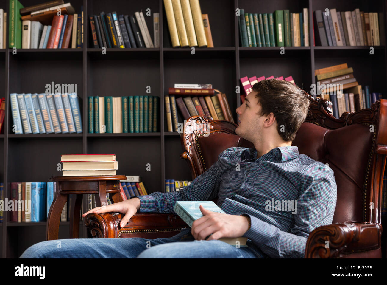 Junger Mann Auschecken Bücher in einer Bibliothek sitzen in einem bequemen Sessel, liest die Titel der Bücher in den Regalen Stockfoto