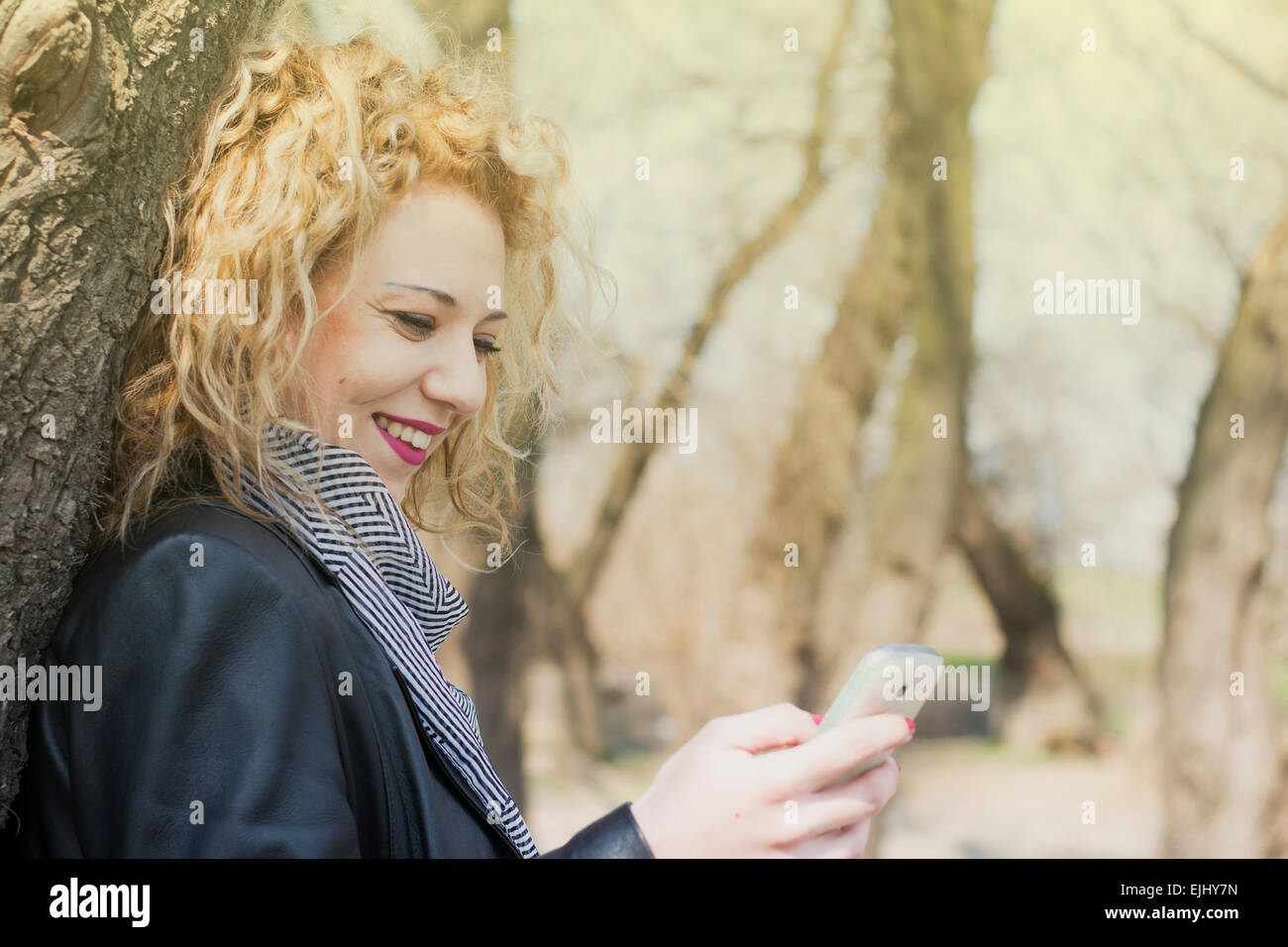 Hübsche junge Frau mit blonden lockigen Haaren auf einen Baum und tippen auf dem Handy. Sie lächelt wie sie mit ihr kommuniziert Stockfoto
