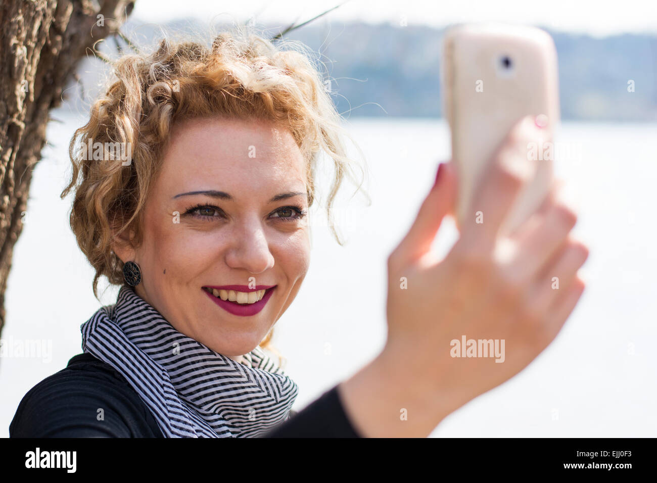 Hübsche junge Frau mit blonden lockigen Haaren nehmen Selfie mit dem Telefon in Natur. Sie lächelt wie sie mit er kommuniziert Stockfoto