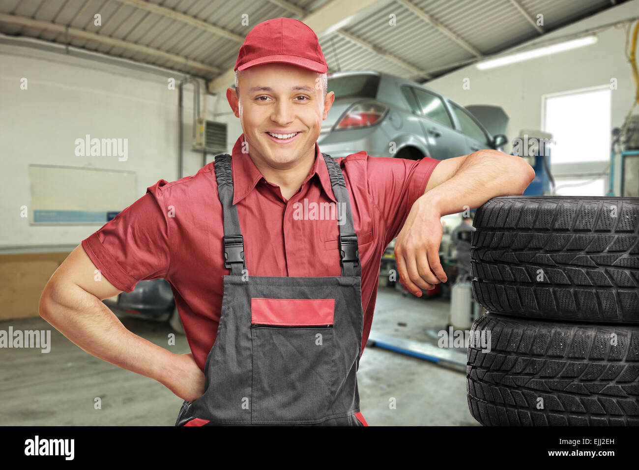 Fröhliche männliche Mechaniker stützte sich auf einen Stapel von Reifen und posiert in einer Garage mit einem Auto hinter ihm Stockfoto