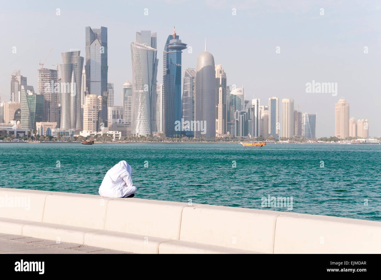 Eine Qatari Mann sitzt allein auf der Stadt am Wasser mit den Wolkenkratzern von Doha im Hintergrund. Stockfoto