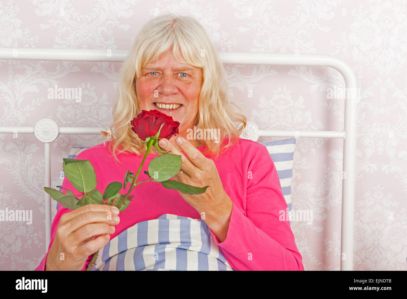 Lächelnde Frau in rosa Pyjama sitzen im Bett mit einer roten rose Stockfoto