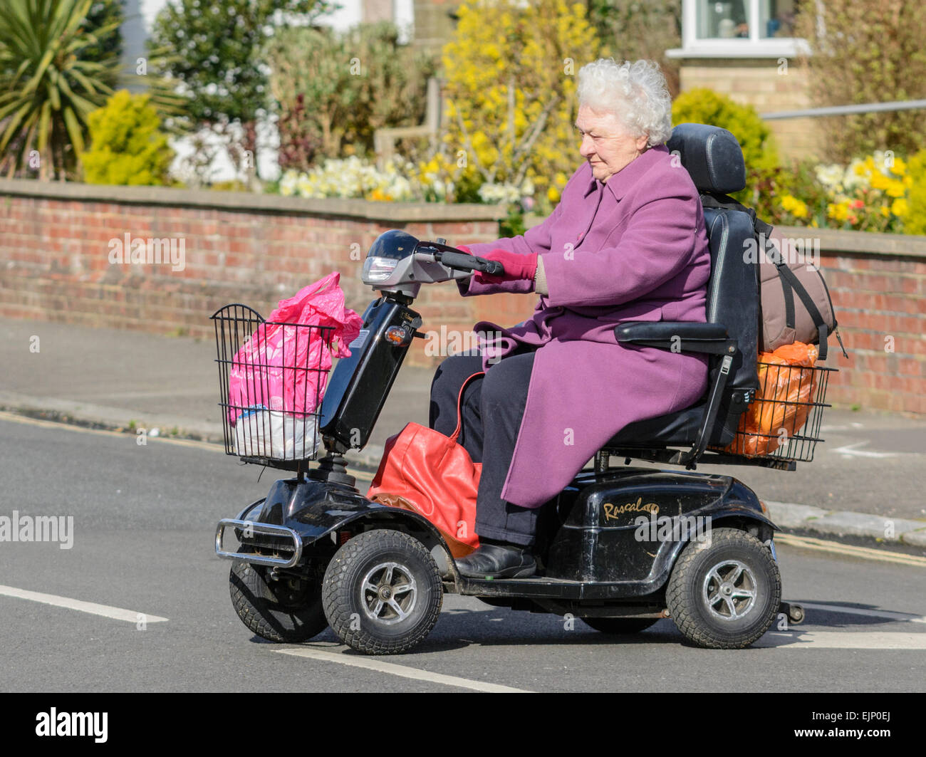 Mobility Scooter. Ältere Frau reiten ein Mobility Scooter über eine Straße, die einkaufstaschen in England, Großbritannien. Stockfoto