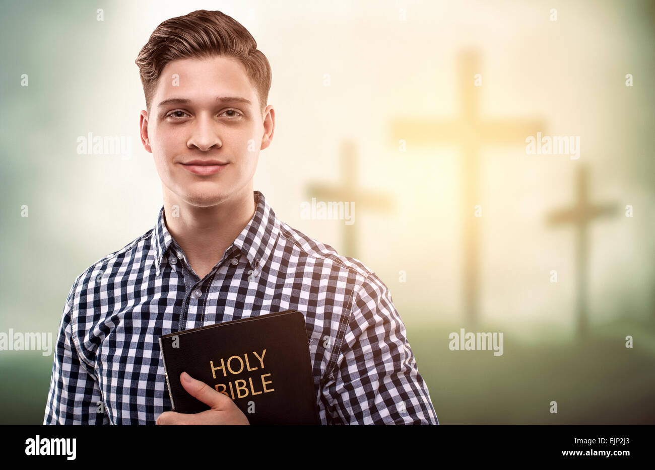 Schöner junger glücklicher Mann hält eine Bibel mit Baum kreuzt Hintergrund (Ostern-Konzept) Stockfoto