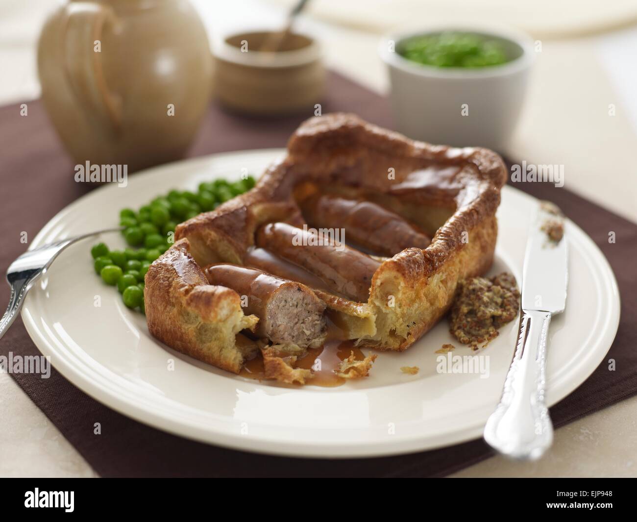 vergoldete Mahlzeit Kröte im Loch große Quadrat Yorkshire Pudding mit drei Würstchen Soße Erbsen und Vollkorn-Senf Stockfoto