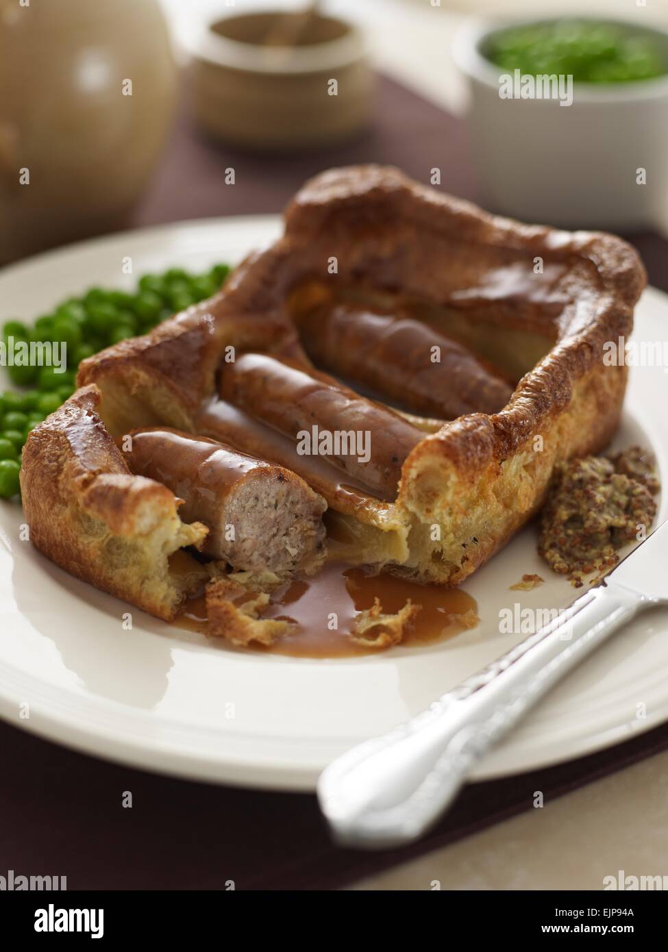 vergoldete Mahlzeit der Kröte im Loch große quadratische Yorkshire Pudding mit drei Würstchen Soße Erbsen und Vollkorn-Senf Stockfoto