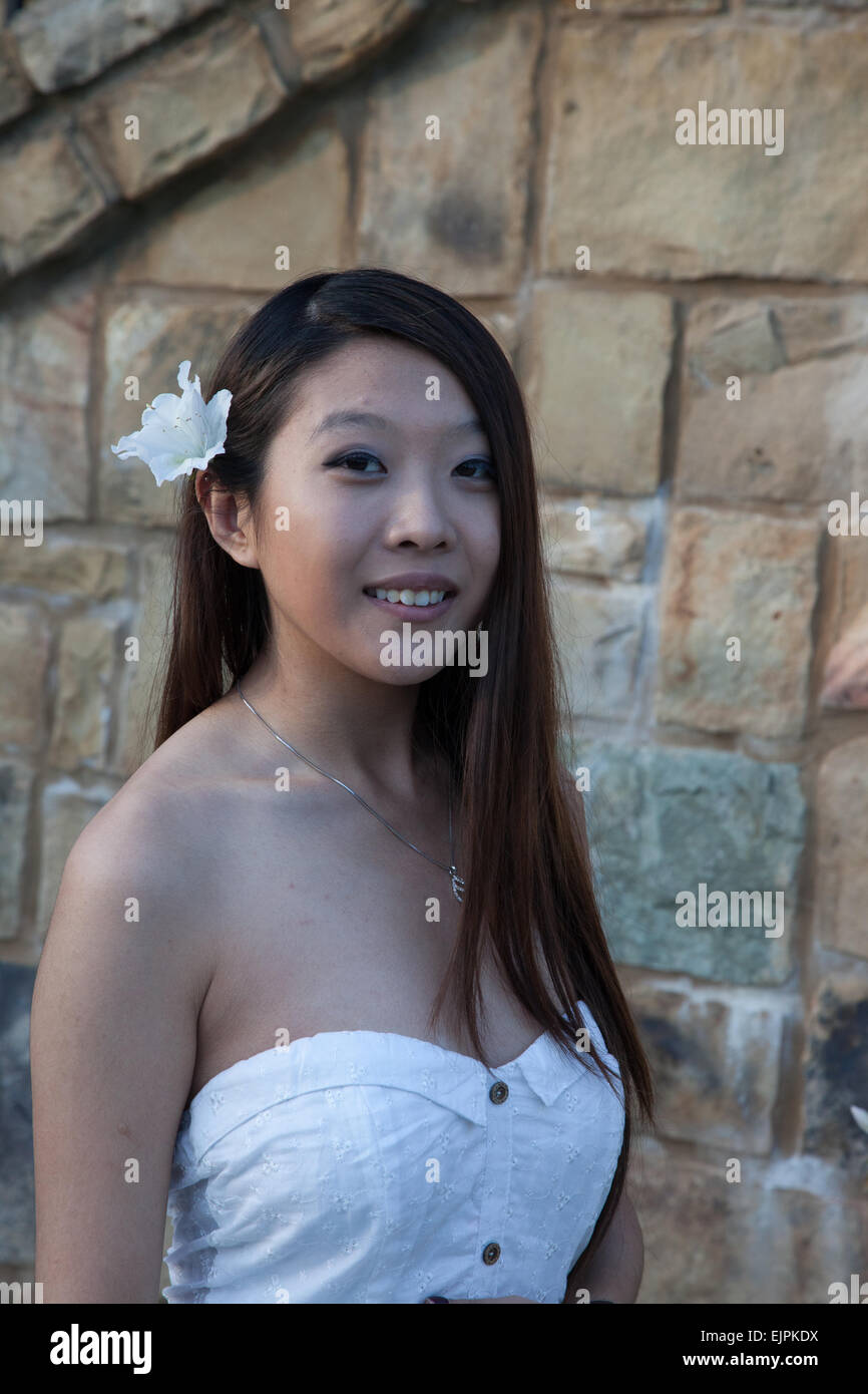 Ziemlich Asian Frau im Freien mit einer steinernen Mauer, in einem weißen Kleid und mit einer weißen Blume im Haar Stockfoto