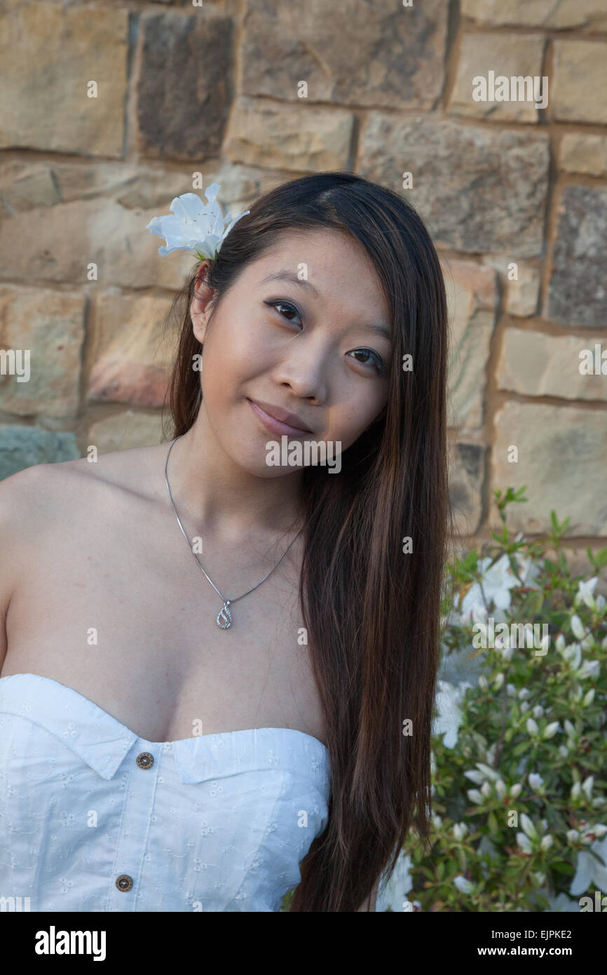 Ziemlich Asian Frau im Freien mit einer steinernen Mauer, in einem weißen Kleid und mit einer weißen Blume im Haar Stockfoto