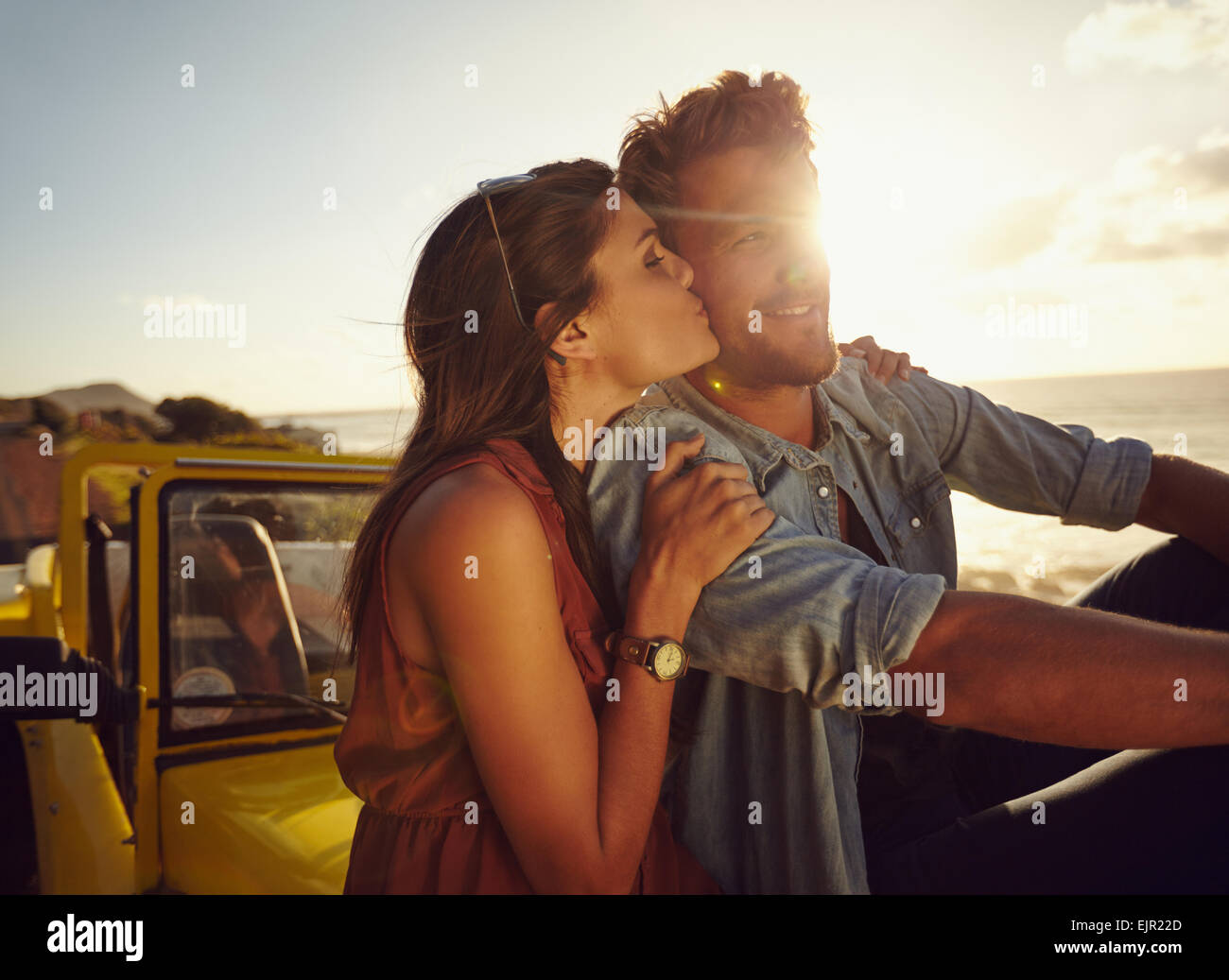 Romantische junges Paar sitzt auf der Motorhaube ihres Autos auf einem Roadtrip. Schöne junge Frau küssen ihren Boyfriend-look Stockfoto