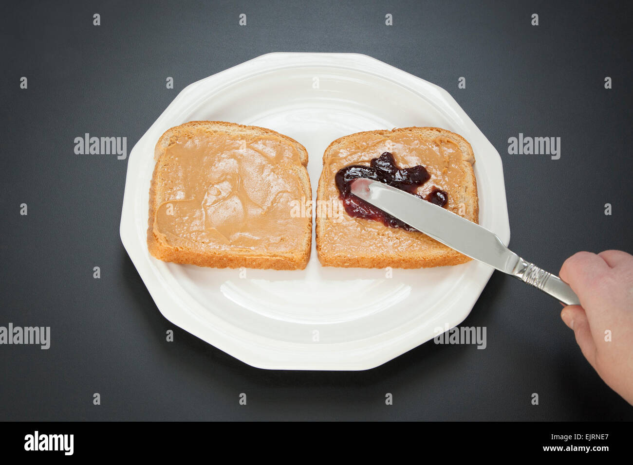 Zwei Scheiben Weißbrot stehen nebeneinander auf einem weißen Teller; Erdnussbutter und Marmelade sind mit einem Messer auf dem Brot verteilen. Stockfoto