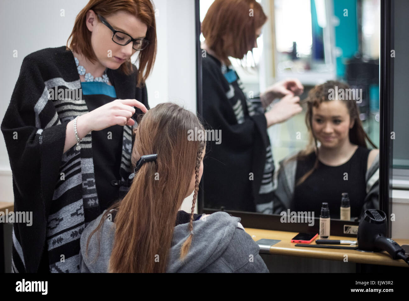 Weibliche Friseur in Gläsern Flechten ein Modell Haar Mode präsentieren sowohl in der Reflexion eines Spiegels in einem Salon. Stockfoto