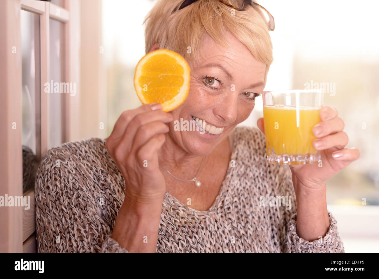 Lachend temperamentvollen blonde Frau mittleren Alters mit einer Orangenscheibe für ihr Auge halten Sie ein Glas frisch gepresster Orangensaft Stockfoto