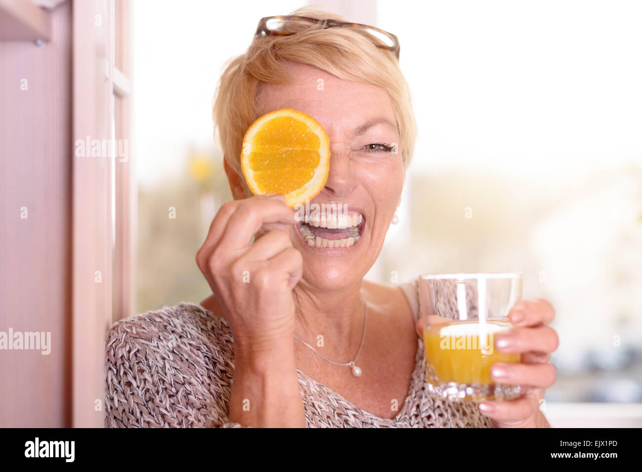 Lachend temperamentvollen blonde Frau mittleren Alters mit einer Orangenscheibe für ihr Auge halten Sie ein Glas frisch gepresster Orangensaft Stockfoto