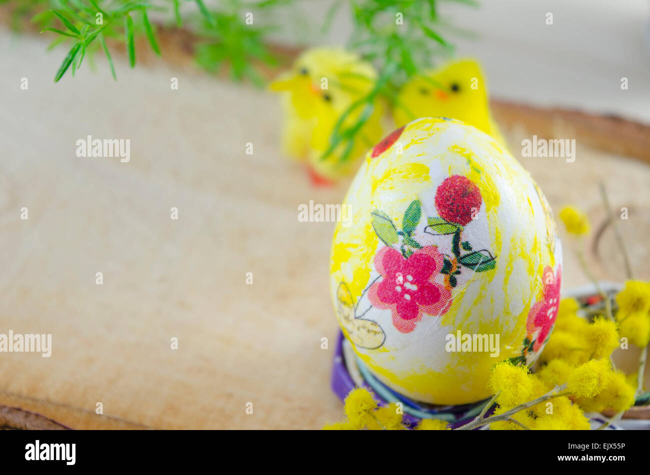 Handgemalte Decoupage Osterei auf einem Ständer mit gelben Blüten und Hühnern im Hintergrund auf einem Holztisch Stockfoto