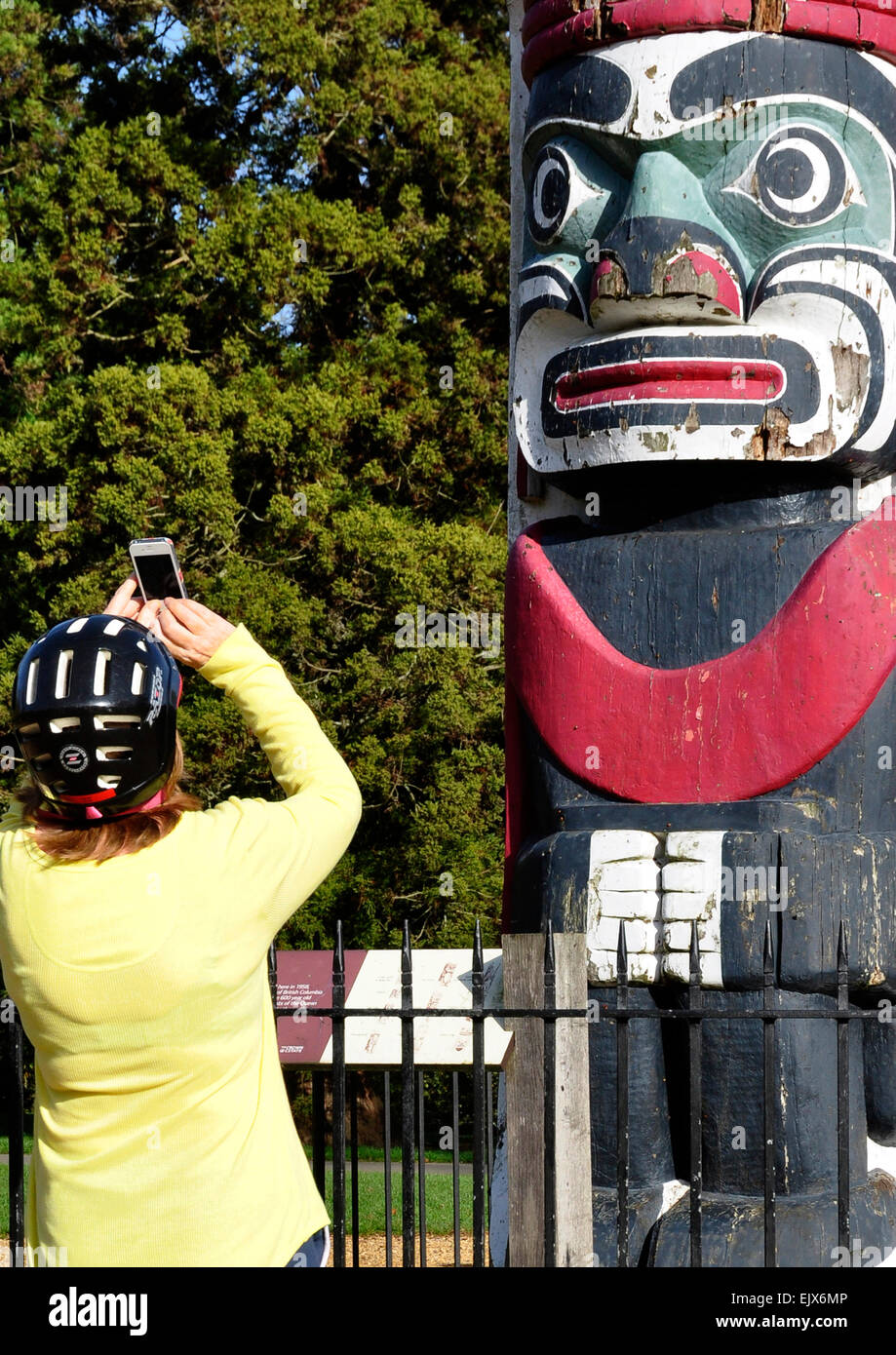 Frau trägt Fahrradhelm - mit digital Phone zu fotografieren geschnitzt Stammes-Gesichts auf hölzernen Totempfahl - Bitte Lächeln!!! Stockfoto