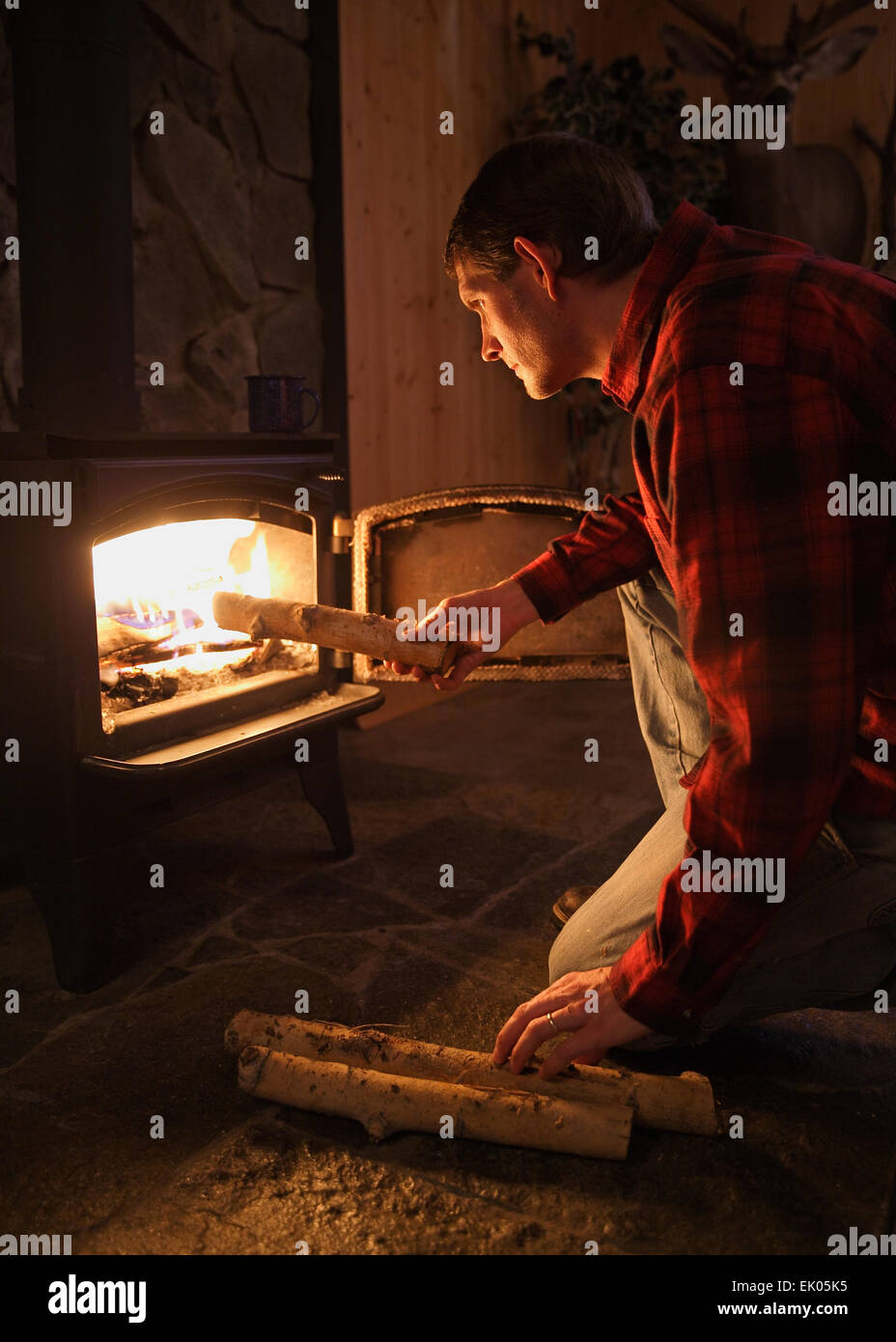Mid-Adult Mann ein Holzbefeuerter Ofen Feuer in rustikaler Umgebung Protokolle hinzufügen Stockfoto
