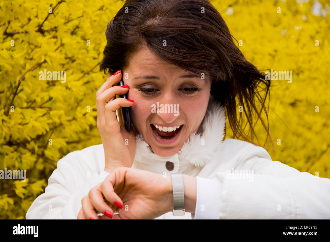 Hübsche junge Frau lächelt während am Telefon zu sprechen und zu realisieren, dass sie spät ist, nachdem sie Armbanduhr sieht. Stockfoto