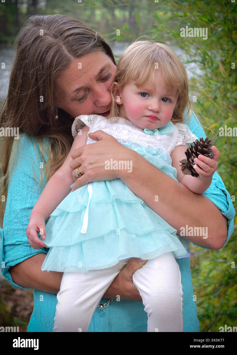 Eine Mutter kuschelt ihr kleines Mädchen, wie sie einen Tannenzapfen an einem See im Stadtpark hält Stockfoto