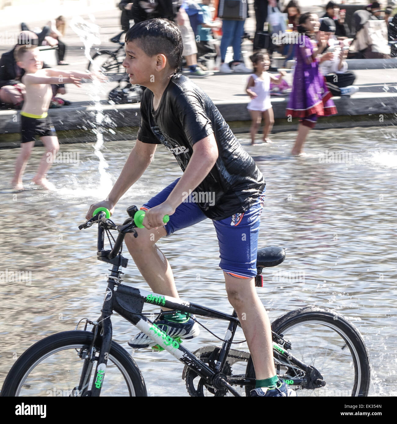 Bradford, West Yorkshire, Großbritannien, 6. April 2015. Erwachsene, Kleinkinder und Jugendliche genießen die Stadtpark mirror Pool und Brunnen im Centenary Square, Großbritanniens größte Stadtzentrum Wasserspiel. Asiatische junge auf Fahrrad im Pool, durchnäßt. Bildnachweis: Mick Flynn/Alamy Live-Nachrichten Stockfoto