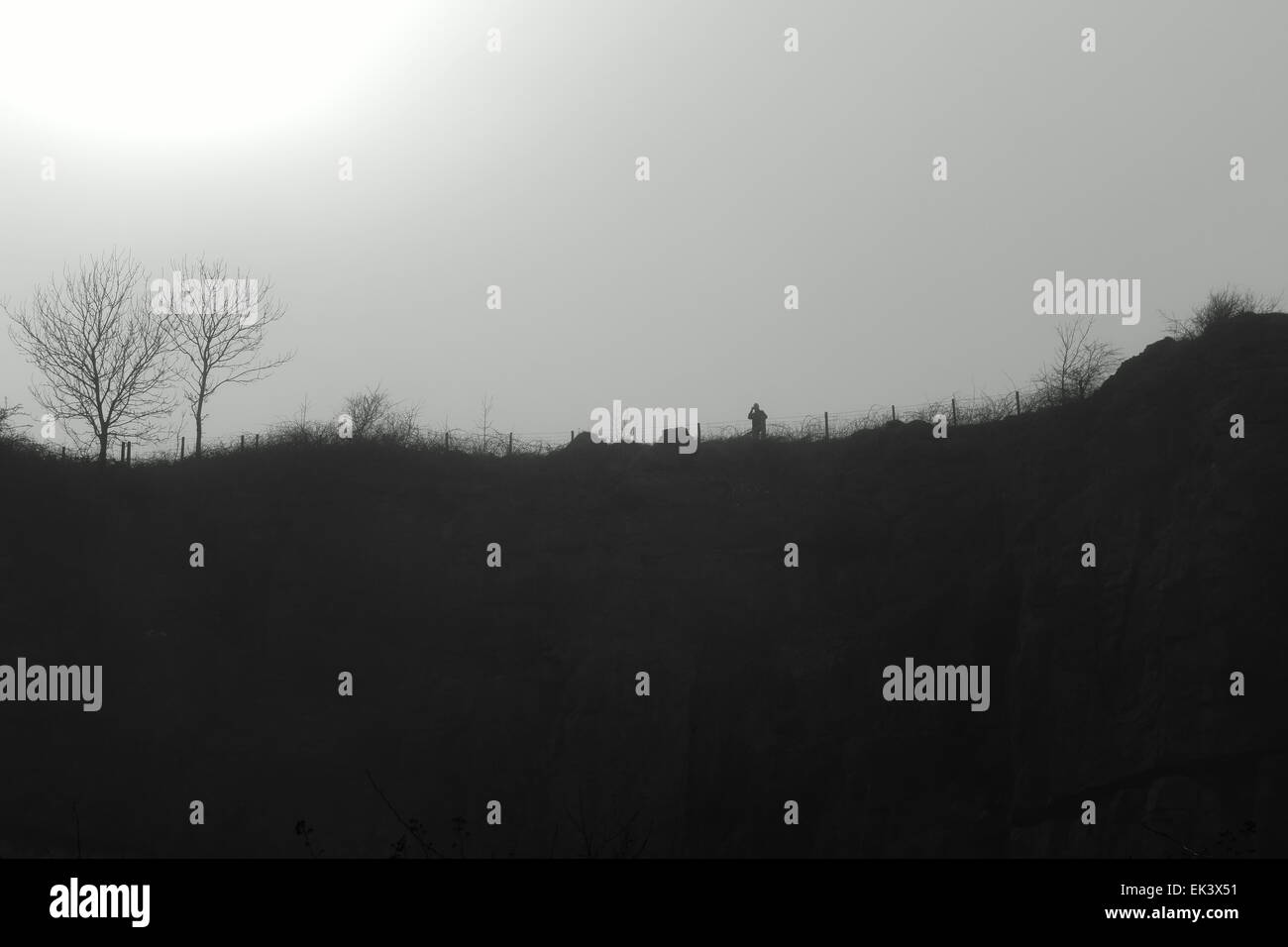 ein Mann schaut von einer Klippe in der Silhouette genommen direkt in die Sonne an einem nebligen Tag in schwarz / weiß Stockfoto