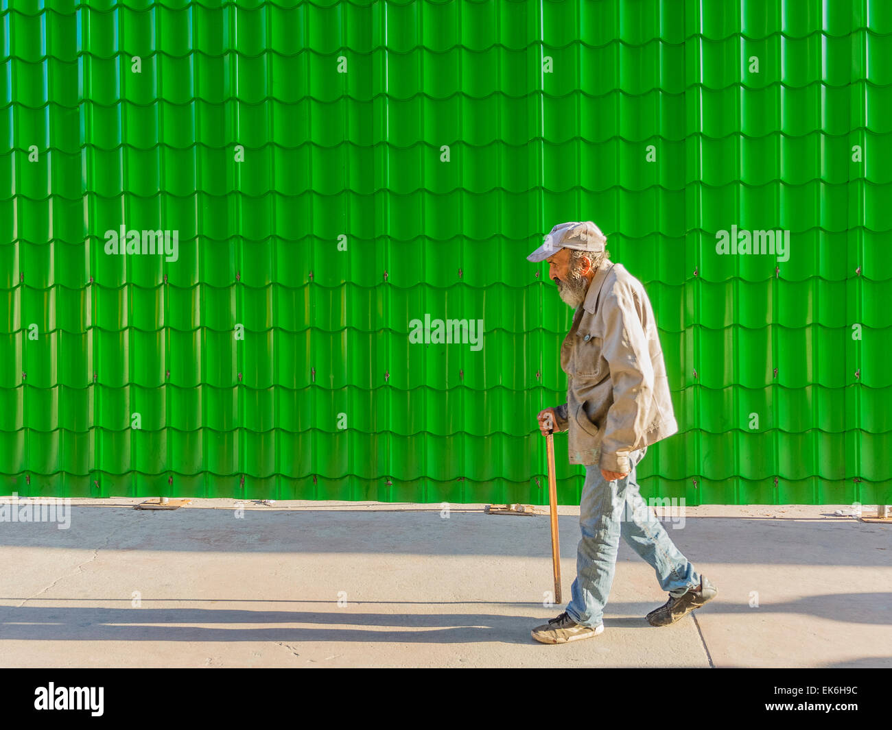 Eine männliche Hispanic Seniorin geht mit einem Stock eine helle grüne Wand aus viele Fliesen nachgeben eine Wiederholung der Form. Stockfoto