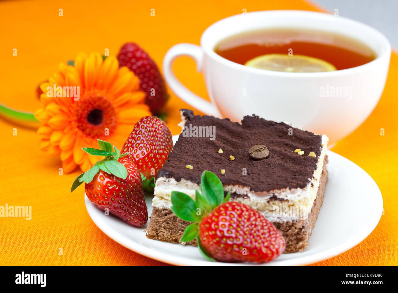 Gerbera, Zitronen-Tee, Kuchen und Erdbeeren liegen auf der orange fa Stockfoto