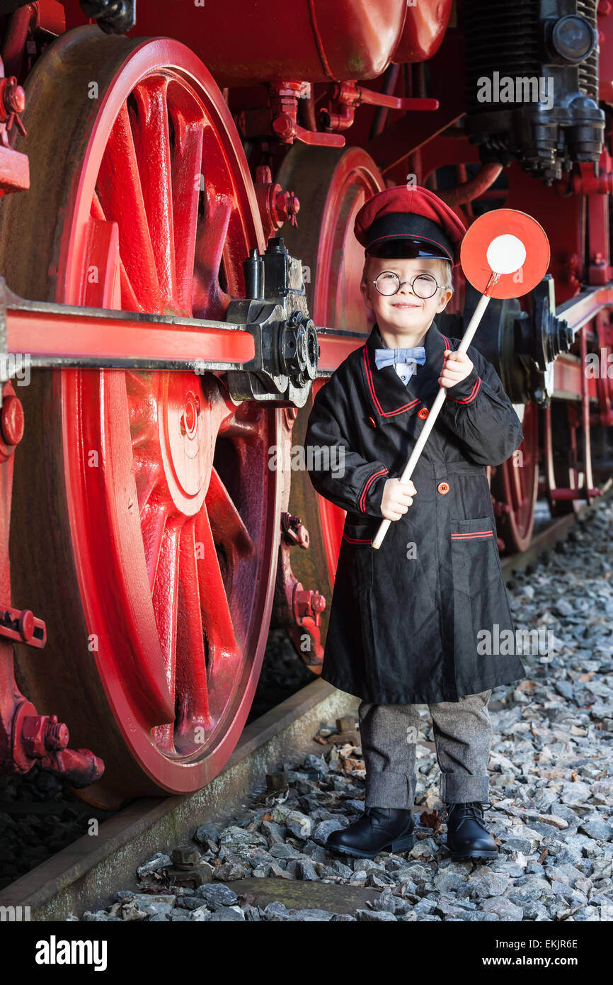 Kleines Kind junge als nostalgische Eisenbahn Leitungsabschnitts mit Kappe und Signalisierung Festplatte neben großen Räder einer Dampflok Stockfoto