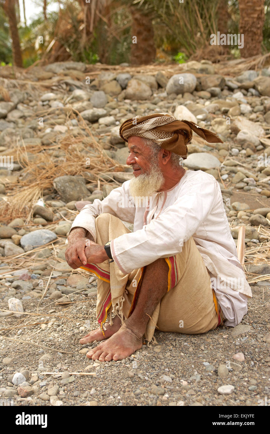 Ein unbekannter omanischer Mann trug ein Muzzar (traditionelle wollenen Turban), entspannt in einem Wadi unterhalb seiner Datum-Plantage. Stockfoto