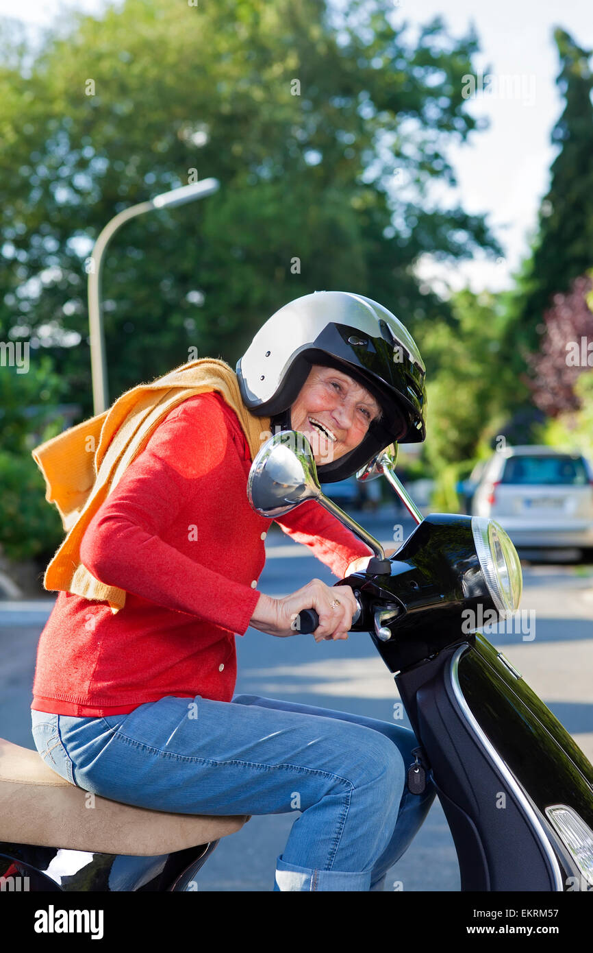 Lebhafte ältere Frau, die einen Motorroller in trendige casual Kleidung gekleidet und trägt eine Sicherheit Helm lachend, als sie, indem ich reitet Stockfoto