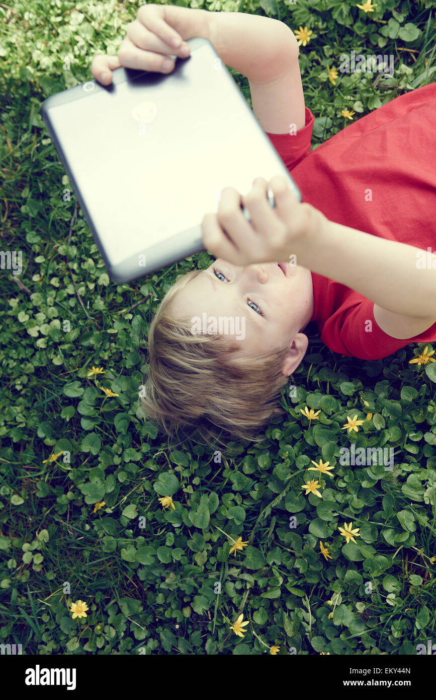 Porträt von Kind blonde junge spielt mit einem digital-Tablette pc Computer im Freien liegen auf Rasen, Gesichtsausdruck Stockfoto