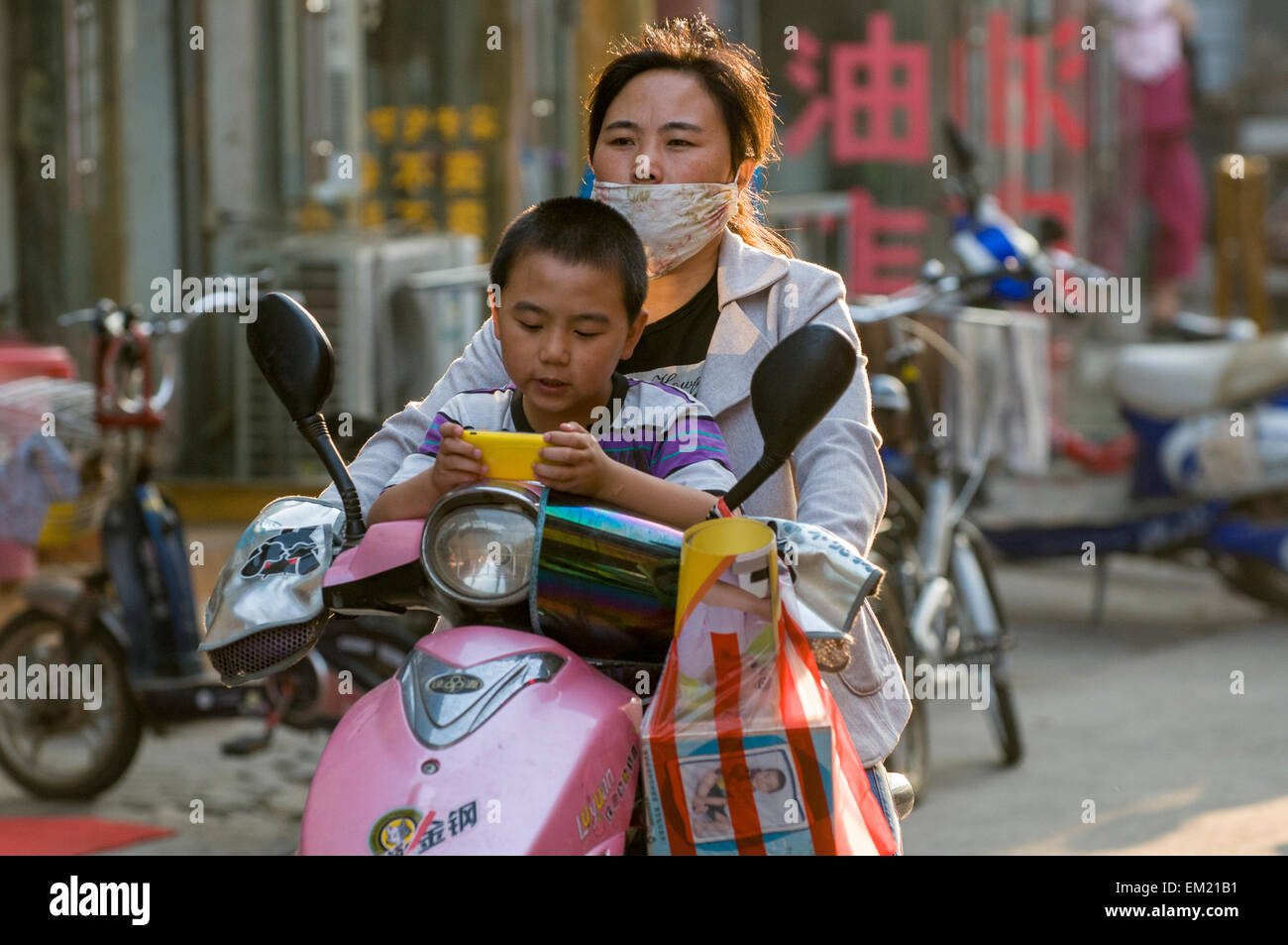 Sohn spielt Videospiel Whle Mutter tragen Gesichtsmaske gegen Verschmutzung Antriebe Scooter, Lianyungang, Provinz Jiansu, China. Stockfoto