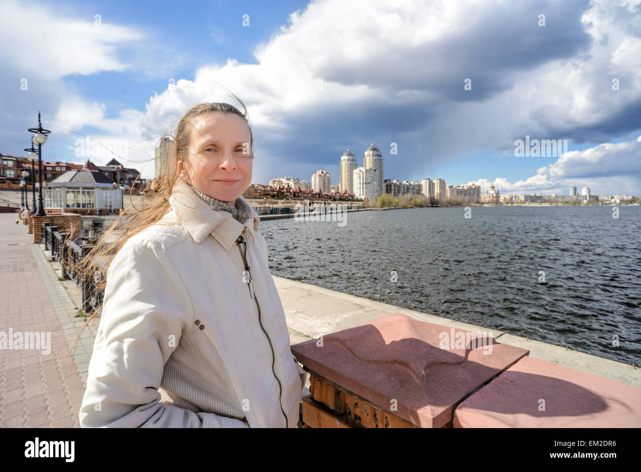 In der Nähe des Flusses steht eine lächelnde Frau, ihr Haar wird durch den Wind unter einem dramatischen stürmischen Frühlingshimmel verschoben Stockfoto