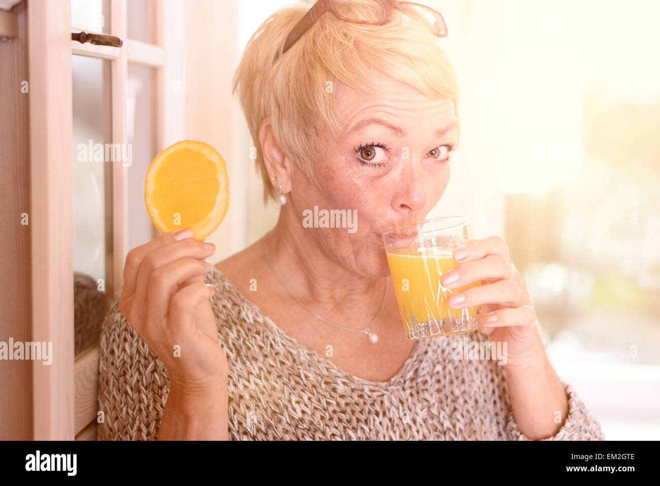 Nahes hohes Mittelalter blonde Frau trinkt Orangensaft und halten eine Orange Fruit Slice während lehnen an der Wand Stockfoto