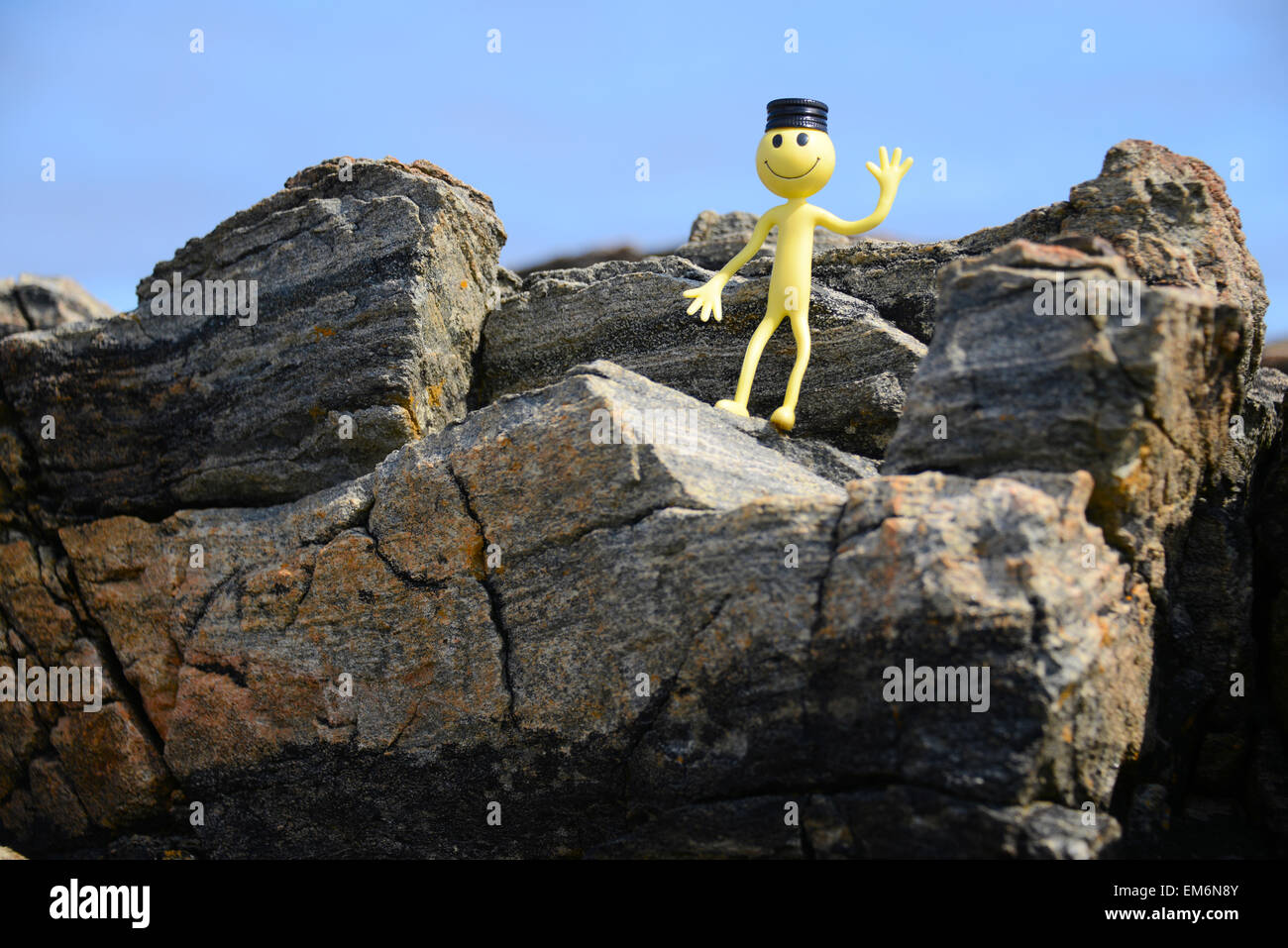 Gelber Smiley-Mann im Urlaub - hier steht er auf einer Klippe trägt eine Weinflasche Top als einen coolen Hut winken Stockfoto