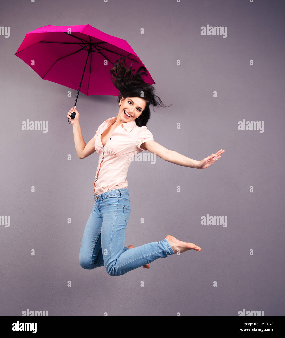 Glückliche junge Frau mit Regenschirm über grauen Hintergrund springen. Blick in die Kamera Stockfoto