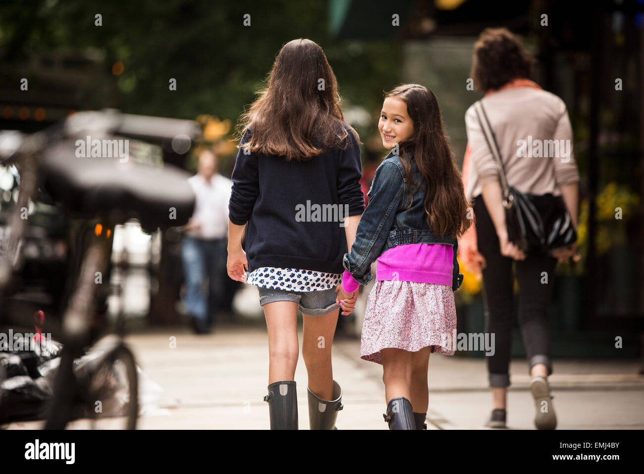 Zwei junge Mädchen Hand in Hand während des Gehens auf Gehsteig, jüngere Mädchen auf der Suche über die Schulter während lächelnd, Rückansicht Stockfoto