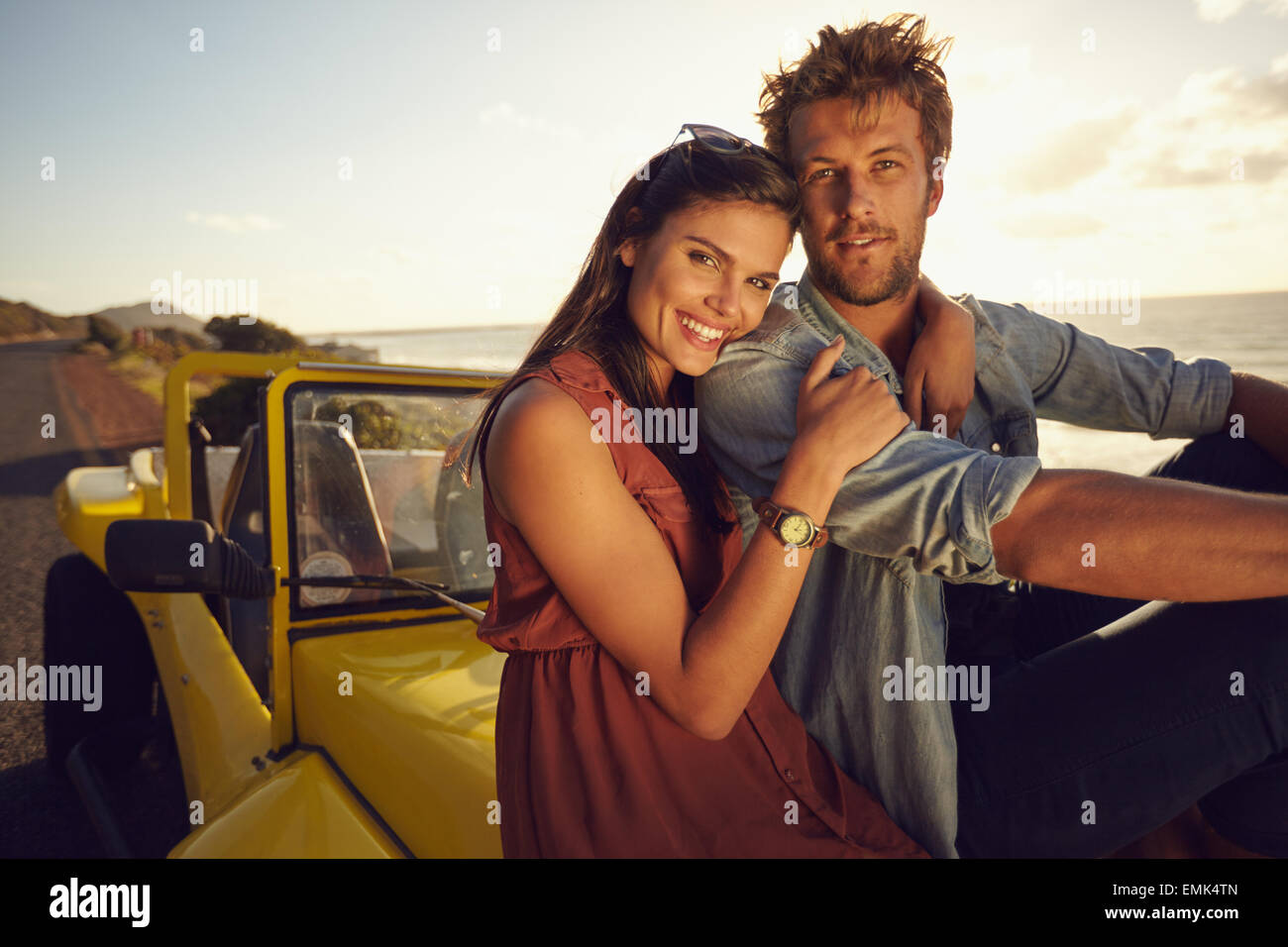 Fröhliches junges Paar sitzt auf der Motorhaube ihres Autos auf einem Roadtrip. Schöne junge Paar zusammen an einem Feiertag. Stockfoto