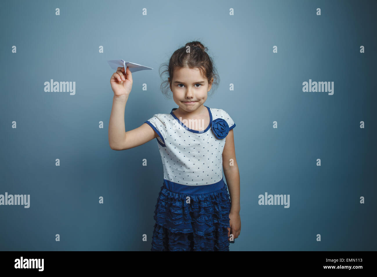eine Mädchen von sieben europäischen Aussehen Brünette hält einen Papierflieger auf einem grauen Hintergrund, Lächeln Stockfoto