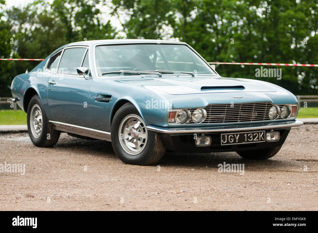 Ein blauer Oldtimer (Aston Martin) aus den frühen 1970er Jahren gezeigt, isoliert, geparkt auf Schotter mit Bäumen hinter. Stockfoto