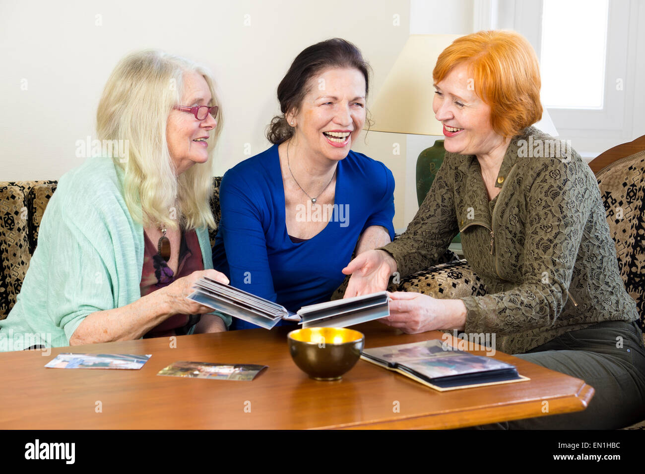 Drei Erwachsene Frauen Freunde entspannen an den lebenden Bereich lachen zusammen während Blick auf ihre alten Fotos in einem Album. Stockfoto