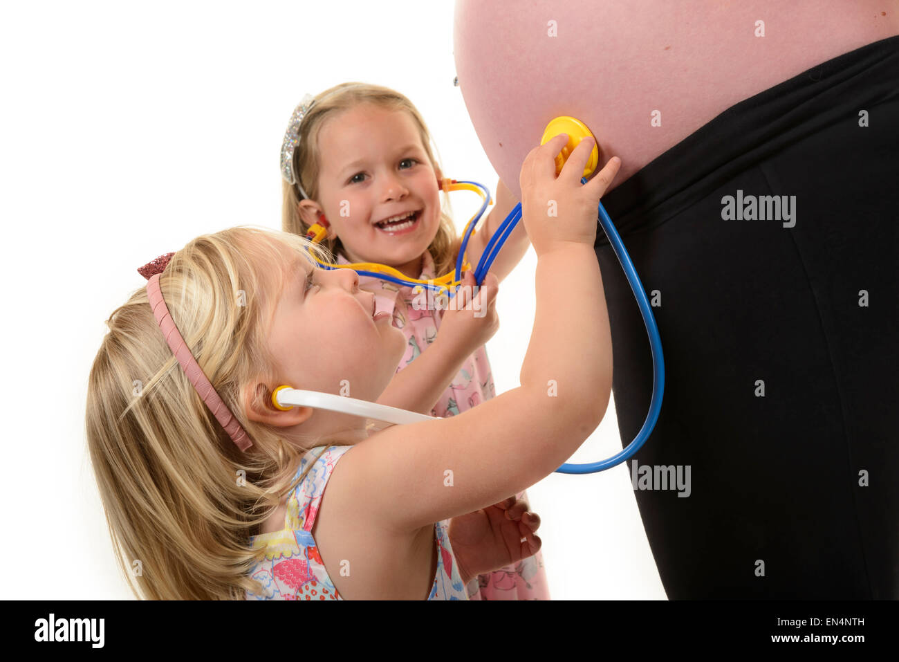 Eine Frau in einem späteren Stadium der Schwangerschaft mit zwei jungen Mädchen Stethoskope an ihrem Baby-Bauch halten. Stockfoto